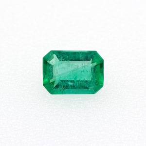 以下が含まれることがあります： 鮮やかな緑色の長方形カットのエメラルド。宝石はファセット加工された表面を持ち、光を反射して透明度を示しています。エメラルドは白い背景に配置され、その色と形を際立たせています。