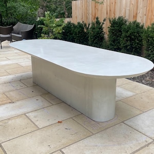 May include: A white oval-shaped concrete outdoor dining table with a smooth, polished surface. The table is on a patio with beige tile flooring.