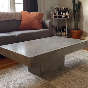 May include: A large, rectangular concrete coffee table with a thick, gray top and a square base. The table is sitting on a light gray rug in a living room.