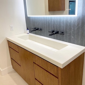 May include: A white countertop bathroom vanity with two sinks and a large mirror above. The vanity has four drawers made of wood with a light brown finish. The countertop is made of a white material that looks like concrete.