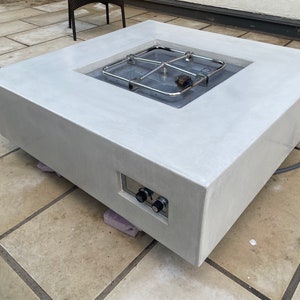 May include: A square concrete fire pit with a stainless steel burner and control knobs. The fire pit is sitting on a patio.