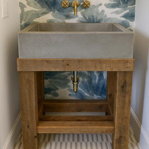 May include: A concrete sink with a wooden stand. The sink is rectangular and has a smooth surface. The stand is made of rustic wood and has a simple design. The sink is set against a wall with a blue and white floral wallpaper.