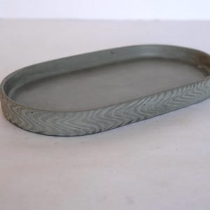 May include: Oval-shaped gray concrete tray with a textured, wave-like pattern around the rim. The tray is a light gray color and sits on a white surface. This decorative tray is suitable for home decor.