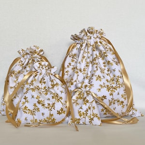 May include: Four drawstring bags of varying sizes. The bags are white with a gold floral pattern and small bee accents. Gold satin ribbon is used for the drawstring closure. The bags are arranged on a white surface.