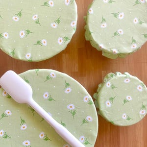 May include: Three round fabric bowl covers in varying sizes featuring a light green background with a white daisy and green leaf pattern, accompanied by a solid white silicone spatula with a long handle and a hole at the end of the handle.