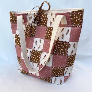 May include: A fabric tote bag with a patchwork design in shades of brown, pink, and white. The bag has cream-colored handles and a brown bow. The squares feature bird and floral patterns. Ideal for carrying items.