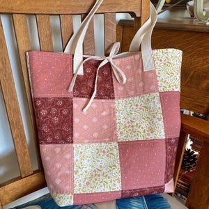 May include: A patchwork tote bag with cream-coloured handles and a matching tie closure. The bag features a variety of pink, burgundy, and floral patterned squares. The bag is sitting on a wooden chair with a blue and white patterned cushion.