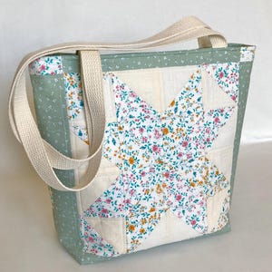 Peut inclure: Un sac fourre-tout matelassé avec un motif d'étoile. Le sac est doté d'une bandoulière crème et est fabriqué avec des patchs de tissu floral et uni dans des tons de vert, crème et rose. Le sac est de forme rectangulaire.