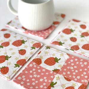 May include: A white ceramic mug rests on a strawberry-patterned coaster. The coasters feature a white background with red strawberries and green leaves, and a pink floral pattern. The coasters are square and quilted.