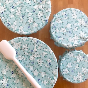 May include: Three round fabric bowl covers in varying sizes featuring a light blue background with a white floral and small blue butterfly pattern, accompanied by a solid white silicone spatula resting diagonally across the largest cover.