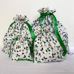 May include: Four white fabric gift bags with a holly berry pattern. The bags are tied with green ribbon. The bags are different sizes, with the largest bag in the back and the smallest in the front.