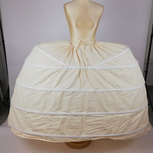 May include: A white, multi-layered petticoat with a gathered skirt and a white ribbon tie at the waist. The petticoat is on a dress form.