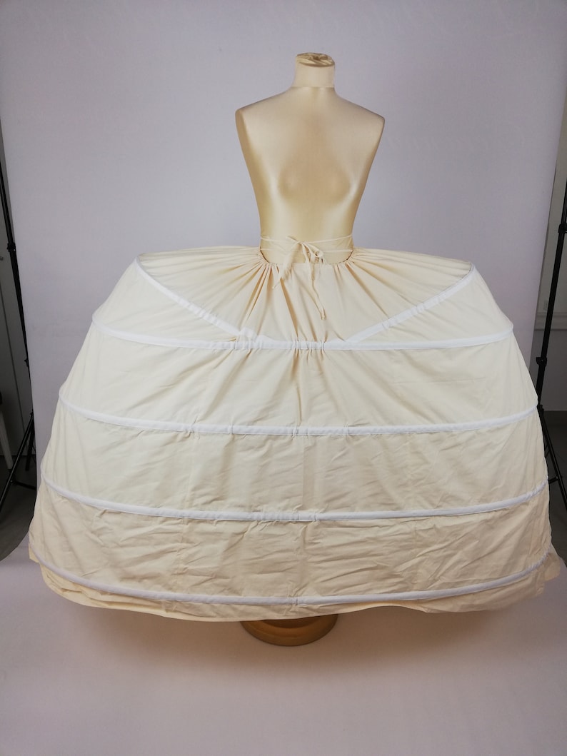May include: A white, multi-layered petticoat with a gathered skirt and a white ribbon tie at the waist. The petticoat is on a dress form.