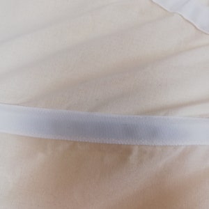 May include: Close-up of a white fabric with a white ribbon sewn along the edge. The fabric has a subtle texture.