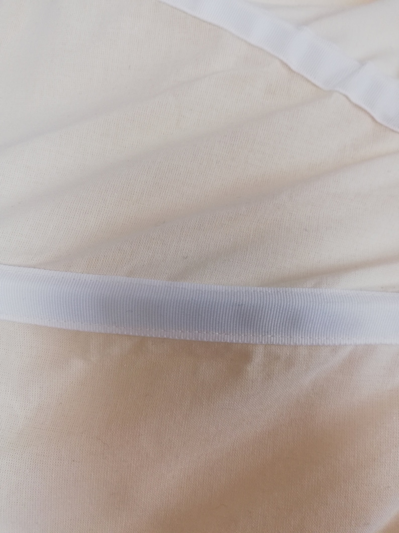 May include: Close-up of a white fabric with a white ribbon sewn along the edge. The fabric has a subtle texture.