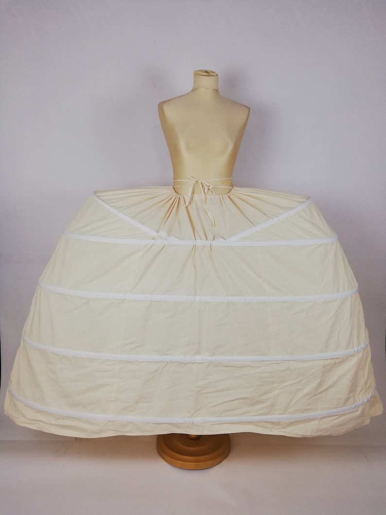 May include: A white petticoat with multiple layers and a gathered skirt. The petticoat is on a dress form and has a white ribbon tied around the waist.
