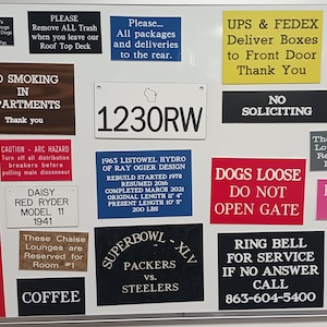 May include: A collection of various rectangular custom engraved signs in multiple colors including red, blue, yellow, black, white, and brown. The signs feature diverse text messages, warnings, and names, displayed together on a white magnetic board.