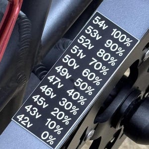 May include: A black and white sticker with voltage and percentage readings. The sticker is mounted on a gray metal frame, with the text "54v 100%" at the top and "42v 0%" at the bottom. The sticker is next to a black gear.