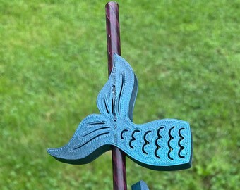 May include: A teal-colored, 3D-printed mermaid tail design on a dark purple and red striped pole. The mermaid tail has a detailed fin and scale pattern. The background is blurred green grass.