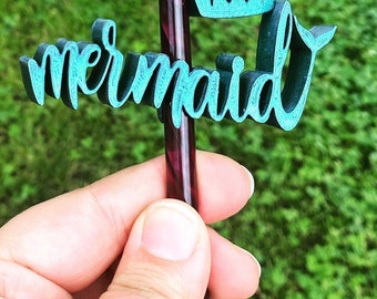 May include: A teal-colored wooden cake topper with the word "mermaid" in a cursive font. The topper is attached to a dark purple and red striped stick, set against a blurred green background.