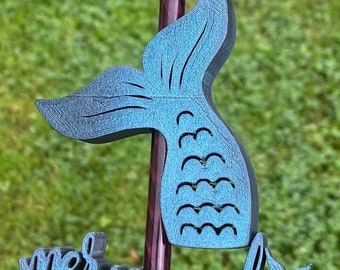 May include: A teal-colored mermaid tail garden stake with a textured finish. The stake is mounted on a purple and brown pole, with the word "mermaid" at the bottom. The background is blurred green grass.