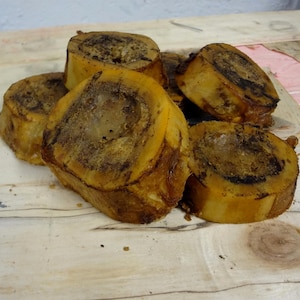May include: A close-up of several large, roasted marrow bones. The bones are a light brown color with dark brown edges and are arranged on a light wood surface.