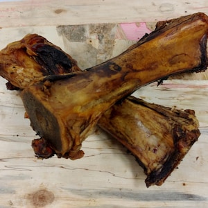 May include: Two large, roasted dog bones with a dark brown, charred exterior and a lighter, tan interior. The bones are positioned diagonally across each other against a light wood grain background.