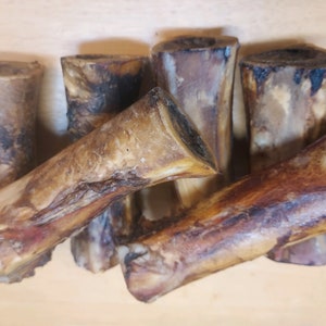 May include: A selection of beef marrow bones, presenting a natural, rustic appearance. The bones exhibit a spectrum of colours, from pale tan to dark brown, with visible textures and variations. The ends are cut, revealing the marrow cavity.