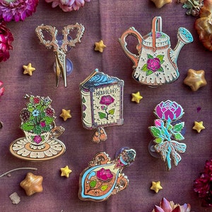 May include: Decorative pins with botanical and garden themes. The collection includes a watering can, teacup with berries, a book labelled "Herbology", a perfume bottle, scissors, and a flower bouquet. The pins are gold-toned with colourful enamel details.