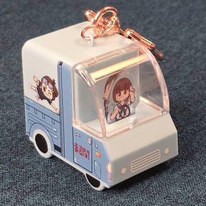 May include: A light blue and white truck-shaped keychain with a clear plastic cab. The keychain features cartoon illustrations of a girl and the Japanese text. The keychain has a rose gold key ring.