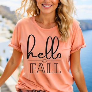 Puede incluir: Una camiseta de color melocotón con el texto "hello fall" en fuente de letra cursiva negra. Un pequeño gráfico de calabaza está por encima de la palabra "fall".