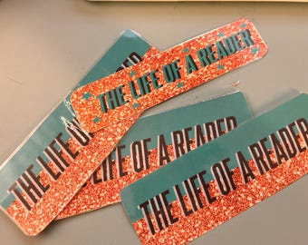 Puede incluir: Marcadores rectangulares con el texto "THE LIFE OF A READER". El texto está en negrita sobre un fondo rojo brillante con detalles y estrellas turquesas. Varios marcadores están apilados.