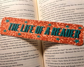 Puede incluir: Marcapáginas naranja brillante con el texto "THE LIFE OF A READER" en turquesa, acentuado con estrellas turquesas. El marcapáginas está en un libro abierto.