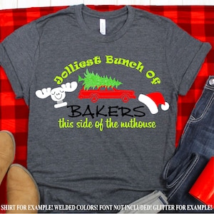 以下が含まれることがあります： お祝いのデザインが施された濃い灰色のTシャツ。「Jolliest Bunch Of BAKERS this side of the nuthouse」というテキストが緑と黒でプリントされています。赤い車が緑のクリスマスツリーを運び、白いヘラジカとサンタの帽子が描かれています。