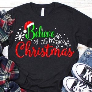 Puede incluir: Una camiseta negra de manga larga con un gráfico verde y rojo que dice "Believe in the Magic of Christmas". El gráfico incluye un gorro de Papá Noel rojo.