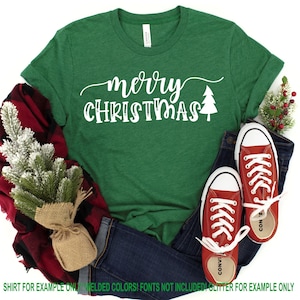 May include: Green t-shirt with the text "merry christmas" in white cursive lettering with a small white Christmas tree graphic. The shirt is folded and displayed on a white background with a red and black plaid blanket, a small potted tree, and a pair of red converse shoes.