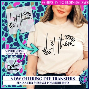 May include: A white t-shirt with the text "Let them" in black script with leopard print accents. The shirt is shown being applied to a t-shirt using a heat press.  The text "Now offering DTF transfers. Send a Etsy message for more info" is at the bottom of the image.
