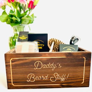 May include: A wooden box with the text "Daddy's Beard Stuff!" engraved on the front. The box contains a variety of beard care products, including a comb, brush, and scissors.