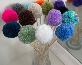 May include: A collection of colorful pom-poms on wooden sticks, arranged in a clear glass vase. The pom-poms are various colors, including pink, blue, green, and purple. The vase sits on a white surface, with a mirror reflecting the arrangement.