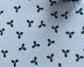 May include: A scattering of green holly leaf cutouts with red berries are spread across a white surface. A dark brown pine cone with glitter accents is in the upper right corner. The image evokes a festive, holiday theme.
