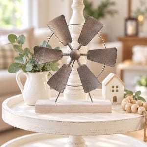 May include: A decorative windmill ornament featuring six weathered metallic gray blades arranged in a circular pattern, mounted on a thin wire frame and supported by a rectangular, light-colored wooden base with a distressed white finish.