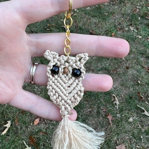 May include: A beige macrame owl keychain with black bead eyes and a long tassel. The keychain is attached to a gold key ring.