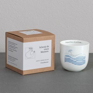 Könnte beinhalten: Eine weiße Porzellantasse mit einer blauen und weißen Illustration eines Segelboots auf Wellen. Die Tasse steht auf einem braunen Karton mit dem aufgedruckten Text "Schenk dir einen Moment. Worte formen Bilder Porzellantasse".