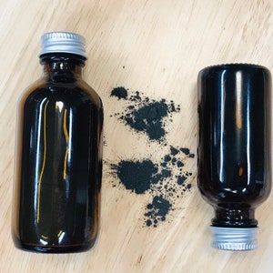 May include: Two amber glass bottles with silver lids, one upright and one lying down, on a wooden surface. Black powder is scattered around the upright bottle.