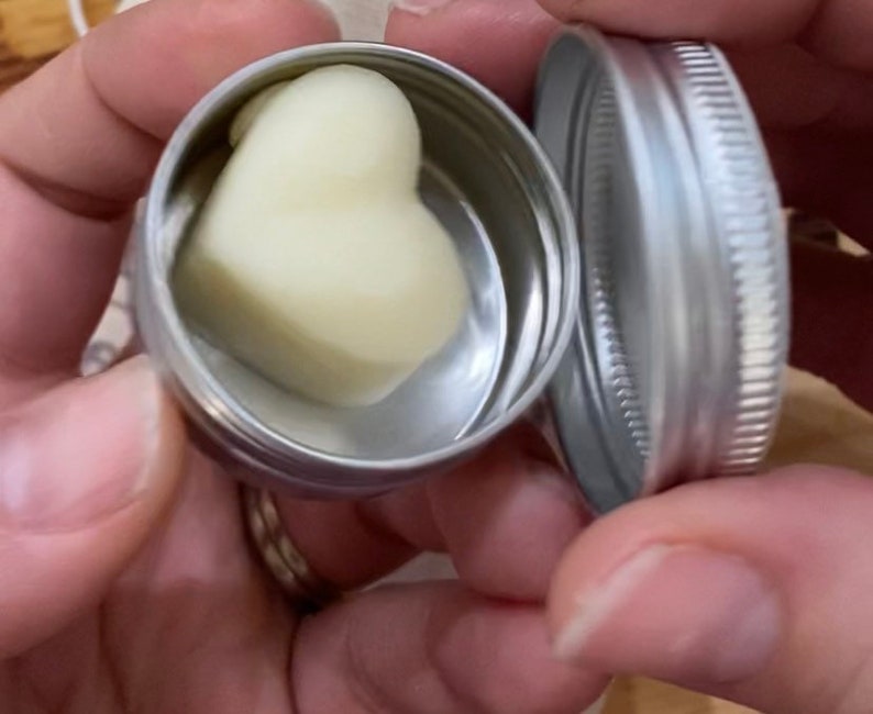 May include: A heart-shaped, white, solid balm in a silver tin with a lid.