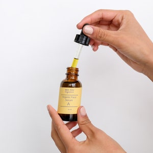 May include: A brown glass bottle with a dropper filled with a yellow liquid. The bottle is labeled "See You Aglow" and "Organic Face Serum A".