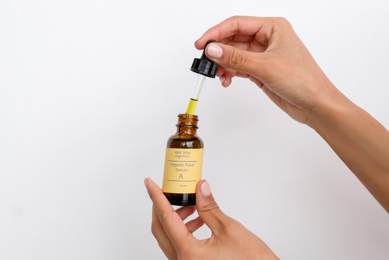 Puede incluir: Una botella de vidrio marr&oacute;n con un gotero lleno de un l&iacute;quido amarillo. La botella est&aacute; etiquetada con "See You Aglow" y "Organic Face Serum A".