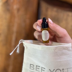 Puede incluir: Un peque&ntilde;o frasco de vidrio marr&oacute;n con un gotero negro en la parte superior, etiquetado como "Bee You Organics", se sostiene en una mano. El frasco est&aacute; en una bolsa de tela blanca con el texto "BEE YOU organics" impreso en ella.