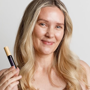 May include: A woman with long blonde hair holds a small brown and purple tube with the word "LASH" printed on it.