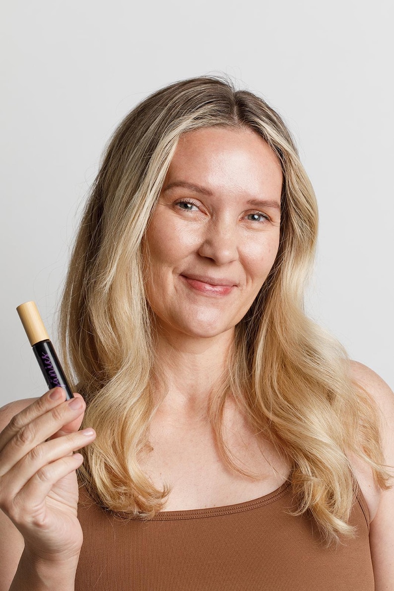 May include: A woman with long blonde hair holds a small brown and purple tube with the word "LASH" printed on it.